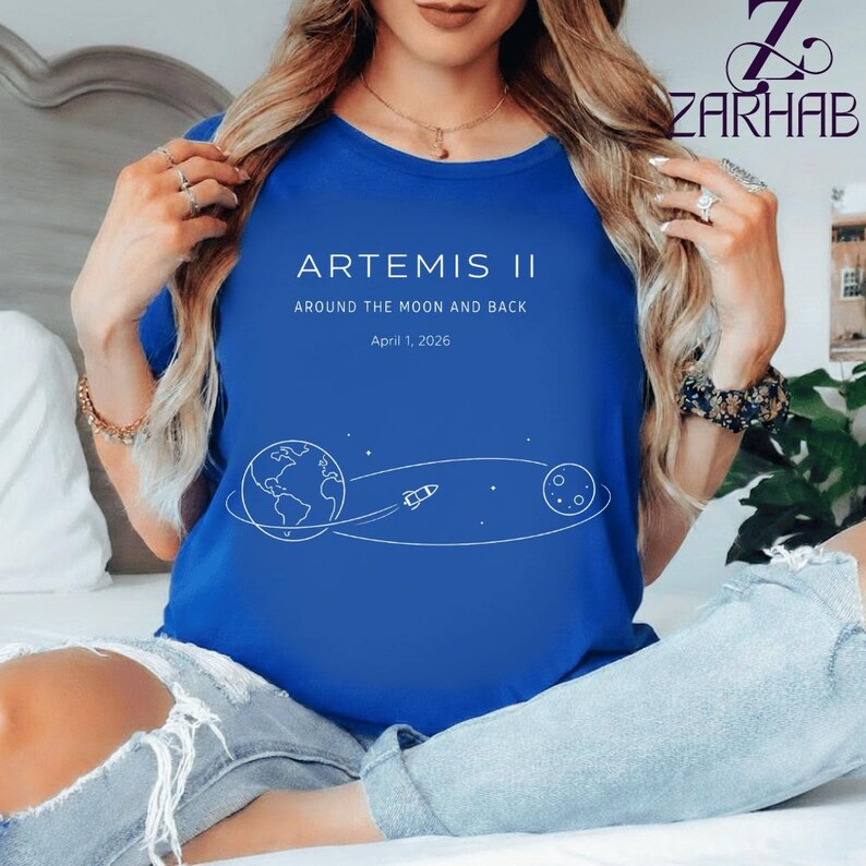 May include: A royal blue t-shirt featuring the text "ARTEMIS II" above the words "AROUND THE MOON AND BACK" and the date "April 1, 2026". A white graphic depicts the Earth, a rocket, and the moon. The shirt is a crew neck.