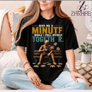 May include: A black short-sleeve t-shirt featuring a graphic of a wooden mannequin sitting on a bench, holding a puzzle piece. The text reads: GIVE ME A MINUTF WHILE I PULL MYSELF TOGETH R. Scattered puzzle pieces are shown on the ground.