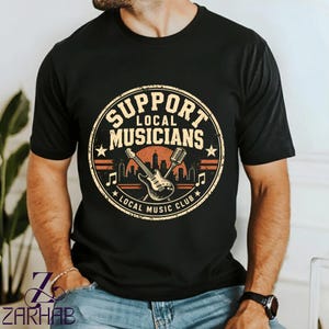 Puede incluir: Una camiseta negra de manga corta con un gráfico circular que incluye el texto "SUPPORT LOCAL MUSICIANS" y "LOCAL MUSIC CLUB". El diseño presenta una guitarra, un micrófono, una silueta urbana y notas musicales en tonos naranja y crema.