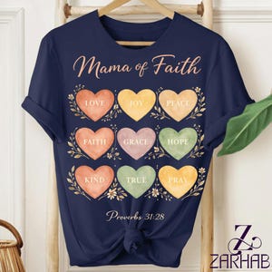 Puede incluir: Camiseta azul marino con el texto "Mama of Faith" y las palabras "Love, Joy, Peace, Faith, Grace, Hope, Kind, True, Pray" dentro de formas de corazón. El texto "Proverbios 31:28" también está impreso en la camiseta.