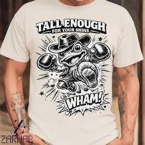 Puede incluir: Camiseta color crema con una rana de dibujos animados en blanco y negro con sombrero de vaquero y guantes de boxeo. El texto "TALL ENOUGH FOR YOUR SHINS" está encima de la rana y "WHAM!" debajo.