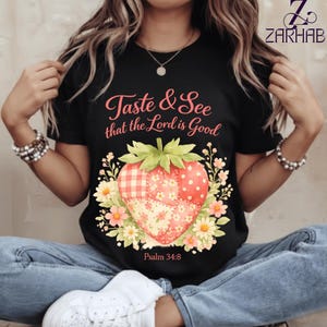 Puede incluir: Camiseta negra con un diseño de fresa. La fresa es un patchwork de lunares rojos, cuadros y estampados florales, rodeada de flores y hojas. El texto "Taste & See that the Lord is Good" está encima de la fresa, con "Salmo 34:8" debajo.