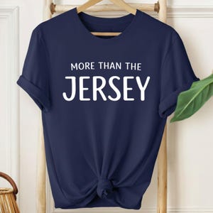 Puede incluir: Camiseta azul marino con el texto blanco "MORE THAN THE JERSEY". La camiseta de manga corta está anudada a la cintura y cuelga de una percha de madera. La camiseta se muestra sobre un fondo claro.