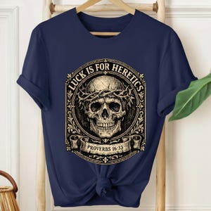 Puede incluir: Camiseta azul marino con una calavera con una corona de espinas, rodeada de adornos decorativos. El texto "LUCK IS FOR HERETICS" está sobre la calavera, con "PROVERBS 16:33" debajo. La camiseta está anudada a la cintura.