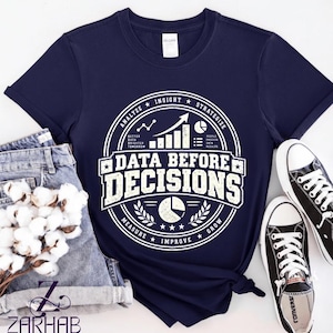 Puede incluir: Una camiseta azul marino de manga corta con un gráfico circular blanco que incluye el texto "DATA BEFORE DECISIONS" y "ANALYZE INSIGHT STRATEGIZE MEASURE IMPROVE GROW" junto con iconos de gráfico de barras, lineal y circular.
