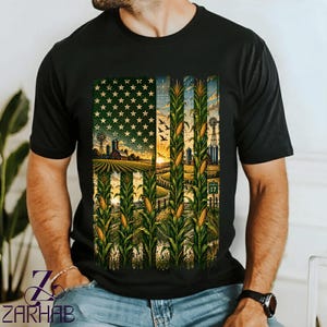 Op de afbeelding: Een zwart t-shirt met een grafische print van de Amerikaanse vlag, samengesteld uit maïsstengels, landbouwvelden en een zonsondergang. Het ontwerp bevat rijen maïs, een schuur, een windmolen en een klein groen verkeersbord met het getal 17.