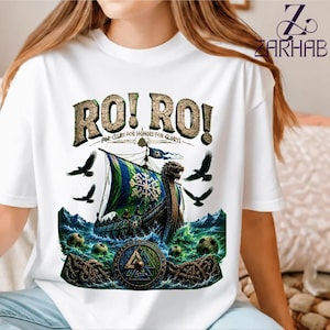 Puede incluir: Una camiseta blanca con un estampado gráfico de un barco vikingo navegando sobre olas turbulentas verdes y azules. El diseño incluye el texto RO! RO! FOR CLUBS FOR HONOR FOR GLORY!, cuervos negros, balones de fútbol y un emblema de nudo nórdico.