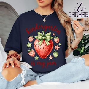 Puede incluir: Una camiseta azul marino oscuro con una fresa roja grande con un lazo de cuadros verdes, rodeada de fresas más pequeñas y estrellas. El texto "kindergarten is my jam" está impreso en letras rojas en la parte frontal de la prenda.