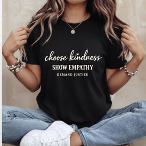 Puede incluir: Camiseta negra con las palabras "choose kindness, show empathy, demand justice" en escritura blanca. La camiseta se lleva con vaqueros azules. La persona lleva pulseras y un collar.