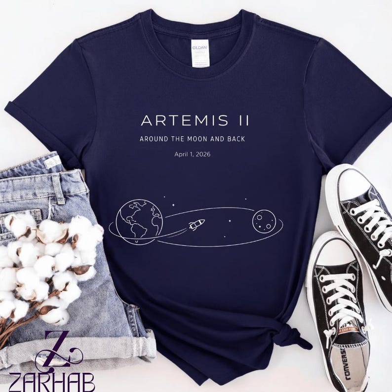 May include: Navy blue t-shirt with white graphic text and illustration. The text reads "ARTEMIS II" and "AROUND THE MOON AND BACK" with the date "April 1, 2026". The graphic shows the Earth, a rocket, and the moon. The shirt is paired with denim shorts and black sneakers.