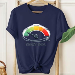 Puede incluir: Camiseta azul marino con un diseño de medidor de nivel de cortisol. El medidor muestra un espectro de colores que va del verde (bajo) al rojo (alto), con la palabra "CORTISOL" impresa debajo. La camiseta está colgada en un perchero de madera.