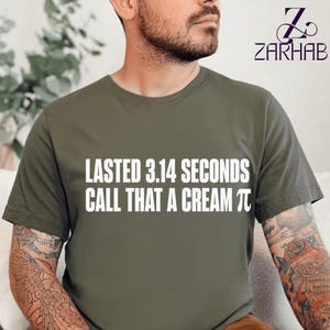 Puede incluir: Una camiseta de manga corta color verde oliva con texto blanco que dice "LASTED 3.14 SECONDS CALL THAT A CREAM PI", donde el símbolo griego pi reemplaza la palabra pi. La prenda tiene cuello redondo y un corte estándar.