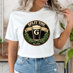 Pode incluir: T-shirt branco com um design circular. O design inclui o texto "SPLIT THE G" acima de um copo de cerveja, uma faixa, trevos e o texto "Est. 1759". A t-shirt é usada com calças de ganga azuis.
