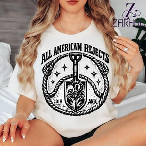 Puede incluir: Camiseta color crema con un diseño gráfico en blanco y negro. El diseño incluye el texto "ALL AMERICAN REJECTS" sobre una pala con un escorpión, rodeado por un borde de cuerda y estrellas. También se incluye el año "2019" y las iniciales "AAR".