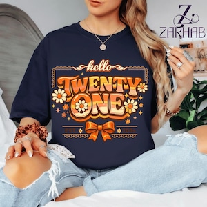 Puede incluir: Camiseta azul marino con el texto "hello TWENTY ONE" en naranja y amarillo, con un diseño retro. El diseño incluye flores y un lazo. También está presente el texto "EST. 2003". Una camiseta informal de estilo vintage.