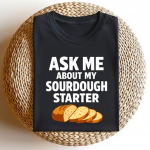 Puede incluir: Camiseta negra con el texto blanco "ASK ME ABOUT MY SOURDOUGH STARTER". Debajo, una ilustración de una hogaza de pan y tres rebanadas. La camiseta se muestra sobre una superficie circular tejida.