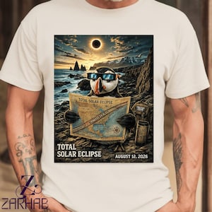 May include: A cream-colored t-shirt featuring a graphic of a puffin wearing eclipse glasses, holding a map labeled TOTAL SOLAR ECLIPSE. The scene includes a solar eclipse in the sky, a rocky coastline, and text reading TOTAL SOLAR ECLIPSE AUGUST 12, 2026.