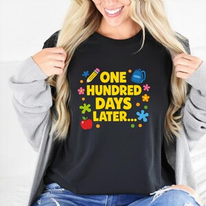 May include: Black t-shirt with the text "ONE HUNDRED DAYS LATER..." in yellow, with colorful graphics of a pencil, backpack, apple, and flowers. The shirt is a crew neck.