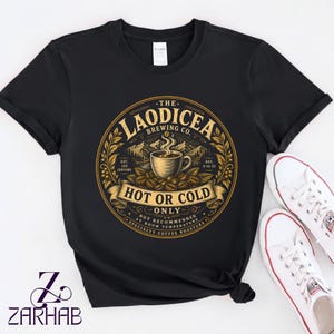 Puede incluir: Una camiseta negra de manga corta con un gráfico circular y texto en dorado y blanco que dice The Laodicea Brewing Co. Hot or Cold Only. El diseño incluye una taza de café humeante, granos de café y montañas en el centro.