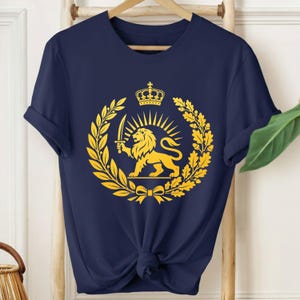 Puede incluir: Camiseta azul marino con un emblema dorado de un león sosteniendo una espada, rodeado por una corona, rayos de sol y hojas de laurel. La camiseta está anudada a la cintura y colgada de una percha de madera.
