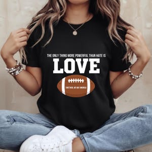 Puede incluir: Camiseta negra con el texto blanco "THE ONLY THING MORE POWERFUL THAN HATE IS LOVE". Debajo, un balón de fútbol americano marrón con cordones blancos y el texto "TOGETHER, WE ARE AMERICA".
