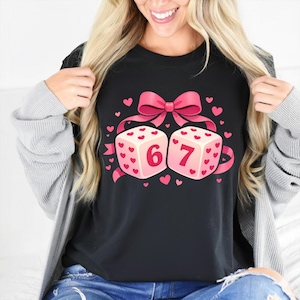 Puede incluir: Camiseta negra con un gráfico de dos dados con los números 6 y 7, adornados con corazones rosas y un lazo rosa. Los dados son de color rosa claro con detalles de corazón rojo. Una cinta rosa conecta los dados y el lazo.