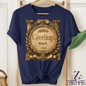 Puede incluir: Una camiseta azul marino con un gráfico dorado de un emblema circular enmarcado por columnas clásicas y hojas de laurel. El texto dentro del emblema dice Greetings. Stop it. —Paul. La camiseta tiene mangas cortas y cuello redondo.