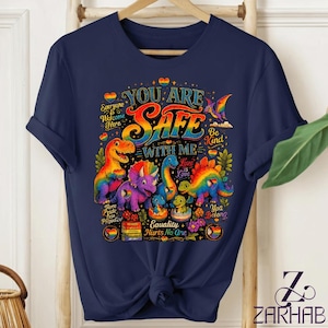 Puede incluir: Una camiseta azul marino con un gráfico colorido de varios dinosaurios y el texto "YOU ARE SAFE WITH ME" junto con "Everyone Is Welcome Here," "Be Kind," "Love Is Love," "Equality Hurts No One," "Pride," y "You Belong."