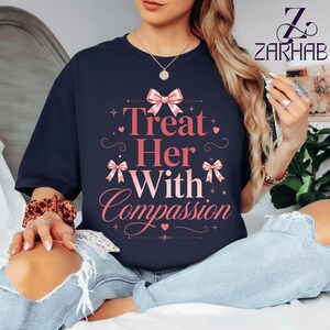 Puede incluir: Camiseta azul marino con el texto rosa "Treat Her With Compassion". El diseño incluye lazos rosas y estrellas. Una prenda de vestir informal.