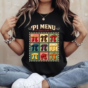 Puede incluir: Camiseta negra con un diseño colorido de "Pi Menu" que presenta porciones de pastel. El diseño incluye el texto "Since 3.14" y varias ilustraciones de pastel en un patrón de cuadrícula. Es una prenda casual.