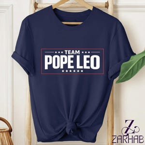 Puede incluir: Camiseta azul marino con el texto blanco "TEAM POPE LEO", enmarcado por un rectángulo rojo y estrellas. La camiseta está anudada en la parte inferior y colgada en un perchero de madera.
