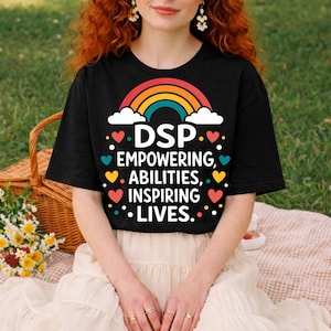 May include: Black t-shirt featuring the text "DSP Empowering Abilities Inspiring Lives" in white, with a rainbow and hearts design. The shirt has a crew neck and short sleeves. The image is taken outdoors.