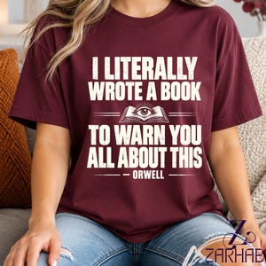 Puede incluir: Una camiseta de manga corta color granate con el texto en blanco I LITERALLY WROTE A BOOK TO WARN YOU ALL ABOUT THIS, un gráfico de un libro abierto con un ojo en el centro y el nombre ORWELL impreso debajo.
