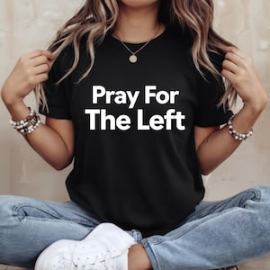 Puede incluir: Camiseta negra con el texto blanco "Pray For The Left". La camiseta es de manga corta y parece estar hecha de un material suave. La persona de la imagen lleva vaqueros azules y zapatillas blancas.