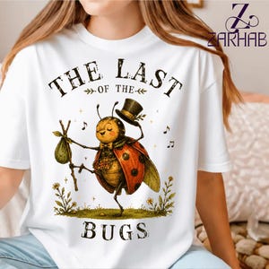 Può includere: Una t-shirt bianca con la grafica di una coccinella che indossa un cilindro e porta un fagotto su un bastone. Il testo recita THE LAST OF THE BUGS. L'illustrazione include piccole note musicali, stelle e fiori su un prato.