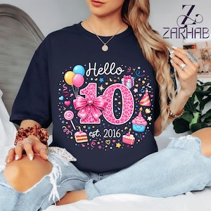 Puede incluir: Camiseta azul marino con un diseño gráfico colorido. El diseño incluye la palabra "Hello", el número "10" con un lazo rosa y el texto "est. 2016". El gráfico también presenta globos, cupcakes y confeti.