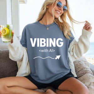 Puede incluir: Una sudadera azul grisáceo con la palabra "VIBING" en letras blancas grandes, con "<with AI>" debajo. Una línea curva blanca con una flecha apunta hacia la derecha. La persona lleva gafas de sol y sostiene una bebida.