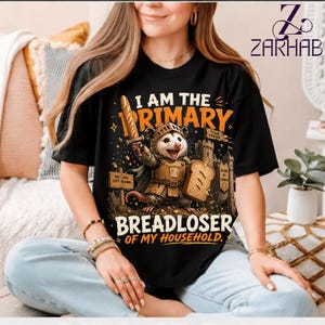 Puede incluir: Una camiseta negra de manga corta con el gráfico de una zarigüeya con armadura medieval, sosteniendo una espada de baguette y un escudo de rebanada de pan. El texto dice I AM THE PRIMARY BREADLOSER OF MY HOUSEHOLD con letreros adicionales.