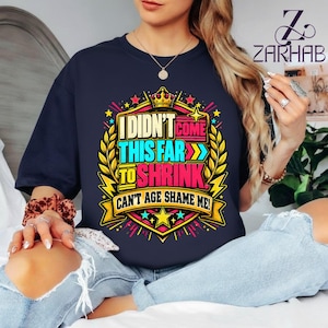 May include: A dark navy blue short-sleeve t-shirt featuring a colourful graphic design with a crown and laurel leaves. The text reads: I DIDN'T COME THIS FAR TO SHRINK. CAN'T AGE SHAME ME! The design uses bright yellow, pink, and light blue accents.