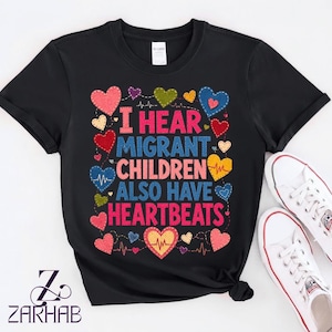 Puede incluir: Una camiseta negra de manga corta con el texto I HEAR MIGRANT CHILDREN ALSO HAVE HEARTBEATS en letras de bloque rosas y azules. El diseño está rodeado por varias formas de corazón coloridas y patrones de líneas de EKG en rosa, azul, amarillo y verde.