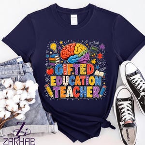 Puede incluir: Una camiseta azul marino de manga corta con un gráfico colorido de un cerebro, libros, una manzana, una calculadora y lápices. El texto GIFTED EDUCATION TEACHER aparece en letras mayúsculas multicolores rodeado de estrellas y corazones.