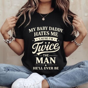 Puede incluir: Camiseta negra con texto blanco: "MY BABY DADDY HATES ME 'CAUSE I'M Twice THE MAN HE'LL EVER BE." La camiseta tiene cuello redondo y mangas cortas. El texto es en negrita y estilizado.