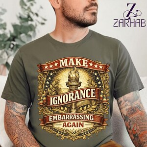Puede incluir: Una camiseta verde oliva con un emblema gráfico en dorado y rojo. El diseño incluye una antorcha y un libro centrales rodeados de filigranas y estrellas. El texto dice "MAKE IGNORANCE EMBARRASSING AGAIN" en letras mayúsculas y negritas.