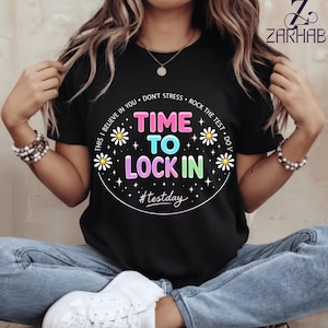 Puede incluir: Camiseta negra con el texto "TIME TO LOCK IN" en rosa, azul y verde, rodeado de margaritas y estrellas. La frase "#testday" está debajo. La camiseta también tiene las frases "THIS I BELIEVE IN YOU", "DON'T STRESS" y "ROCK THE TEST".