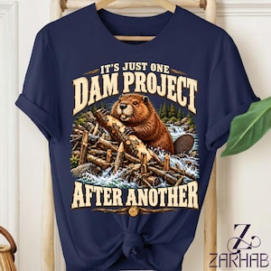 Puede incluir: Una camiseta azul marino con un gráfico de un castor marrón trabajando en una presa de madera en un río. El texto sobre el castor dice IT'S JUST ONE DAM PROJECT y el texto debajo dice AFTER ANOTHER en una fuente clara y llamativa.