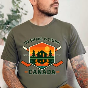 Puede incluir: Camiseta verde oliva con un gráfico que presenta una cabaña, árboles y una puesta de sol sobre el agua. El texto dice "THE COTTAGE IS CALLING CANADA" con palos de hockey y el año 2026.