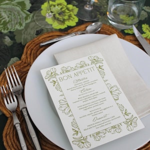 May include: A formal table setting with a white plate, silverware, and a menu. The menu, bordered with floral designs, reads "Bon Appétit" and lists a multi-course meal. A linen napkin and water glass complete the elegant arrangement.