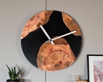 Puede incluir: Reloj de pared redondo de madera con diseño de vetas naturales y detalles de resina negra. El reloj tiene manecillas plateadas y una estética moderna. El diámetro del reloj es de aproximadamente 30 cm, añadiendo un toque rústico.