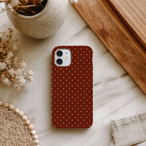 Peut inclure: Une coque de smartphone rouge foncé ornée d'un motif répétitif de petits pois blancs. La coque présente une forme rectangulaire aux coins arrondis avec une découpe circulaire dans le coin supérieur gauche pour l'objectif et le flash.