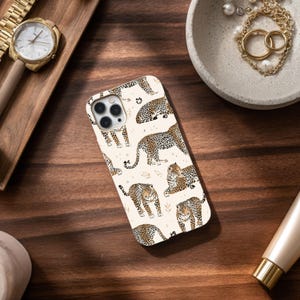 Peut inclure: Une coque de smartphone rectangulaire arborant un motif répétitif de léopards illustrés dans des tons marron, noir et beige sur un fond crème. La coque présente une découpe pour un système de triple appareil photo dans le coin supérieur gauche.